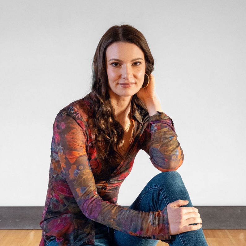 Chelsea Alban, founder of Cosmic Counseling, sitting on the floor in a floral blouse and jeans, embodying her grounded, soulful approach to healing