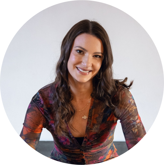 Warm, inviting headshot of Chelsea Alban with shoulder-length brown hair styled in loose waves. She's wearing a semi-sheer, long-sleeved blouse with a dark background and colorful floral print. She is smiling gently and looking directly at the viewer, creating a welcoming and approachable image.
