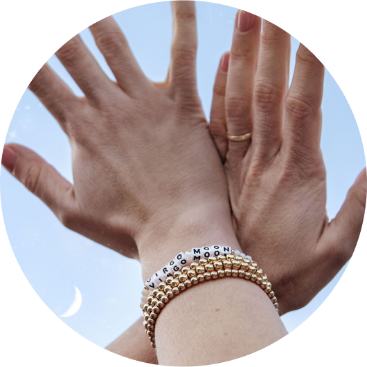 Close-up of a person's hands reaching upward against a light blue sky. They are wearing several gold beaded bracelets with the words "Virgo" and "Moon" spelled out on white beads.