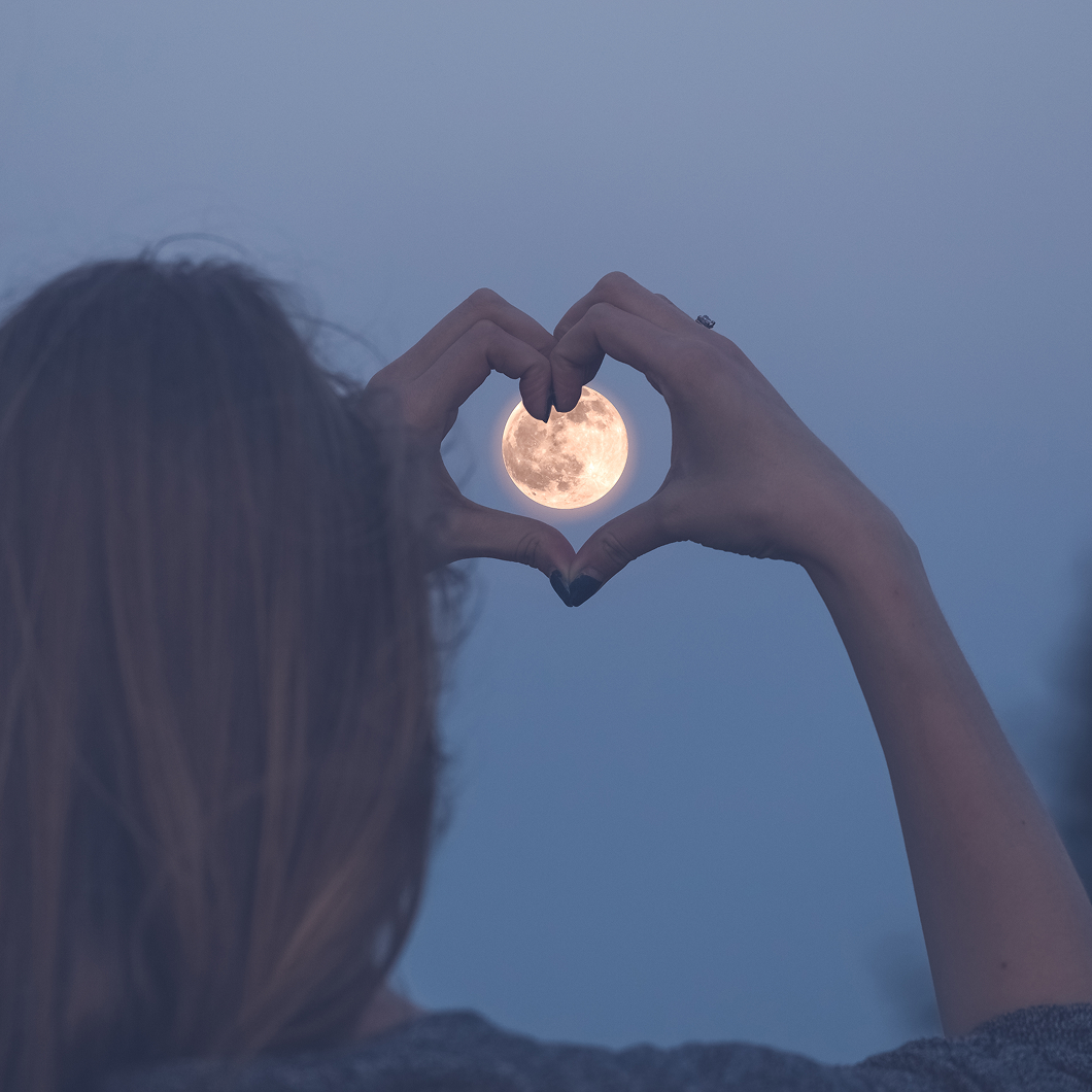 The back of a woman's head is visible as she holds her hands up to form a heart shape, perfectly framing a large, bright full moon in the evening sky.