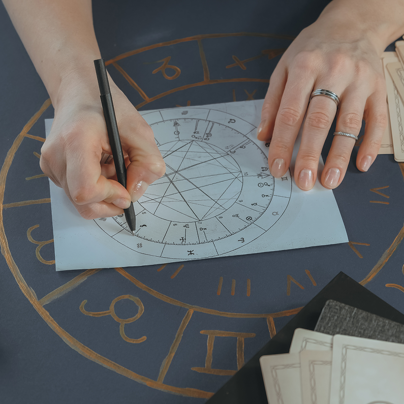 Close-up of hands meticulously drawing lines on a blank astrological birth chart. The chart is laid on a dark surface with a larger, hand-drawn astrological wheel visible in the background. Tarot cards and other tools are also present.