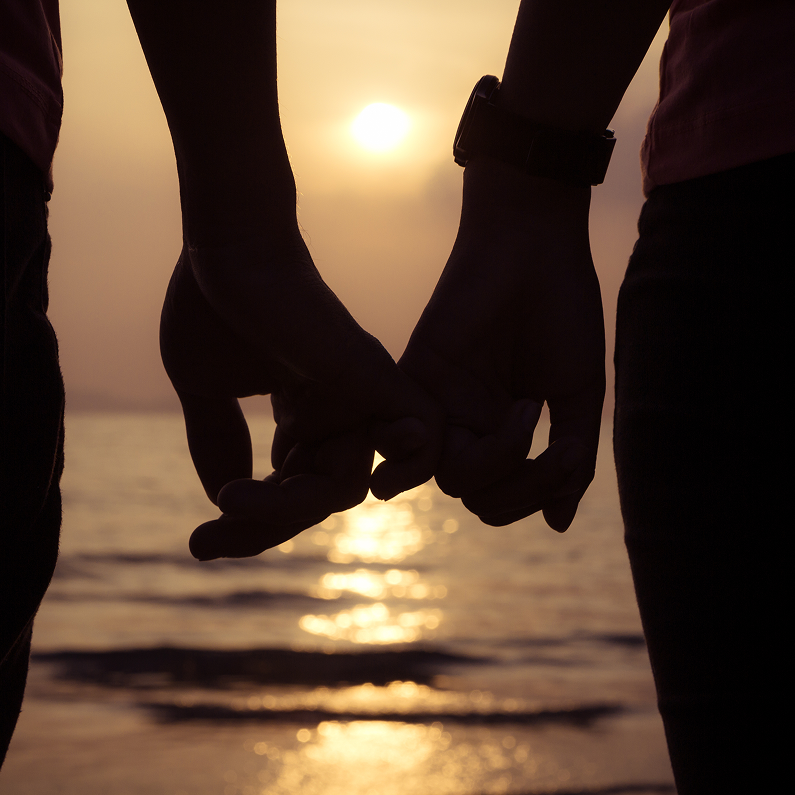 Close-up silhouette of a couple holding hands at sunset, their fingers intertwined. The ocean and setting sun are visible in the background